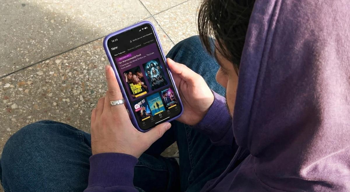 Person in purple hoodie sitting outdoors holding a smartphone with purple case displaying the Galatea app interface with colorful book covers on a concrete background