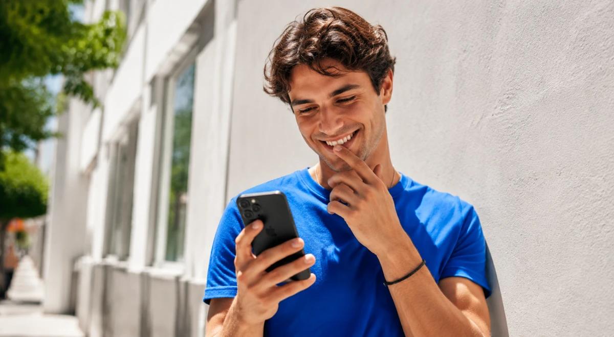 Smiling young man in a blue t-shirt reading a text message on his phone outdoors near a white wall, illustrating how to flirt over text