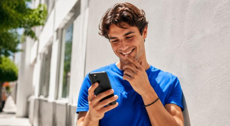 Smiling young man in a blue t-shirt reading a text message on his phone outdoors near a white wall, illustrating how to flirt over text