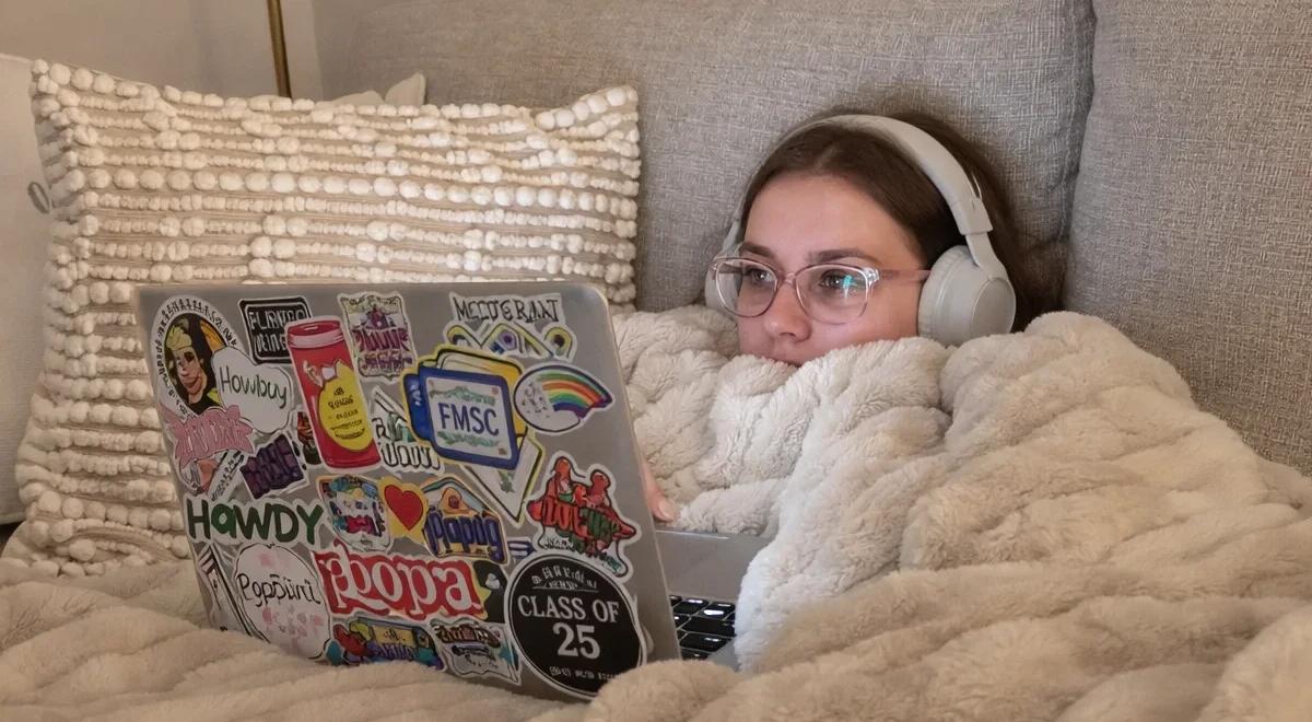 Young woman with headphones and glasses lying on a couch under a fluffy blanket, using a sticker-covered laptop — reflecting cozy digital habits and social media use