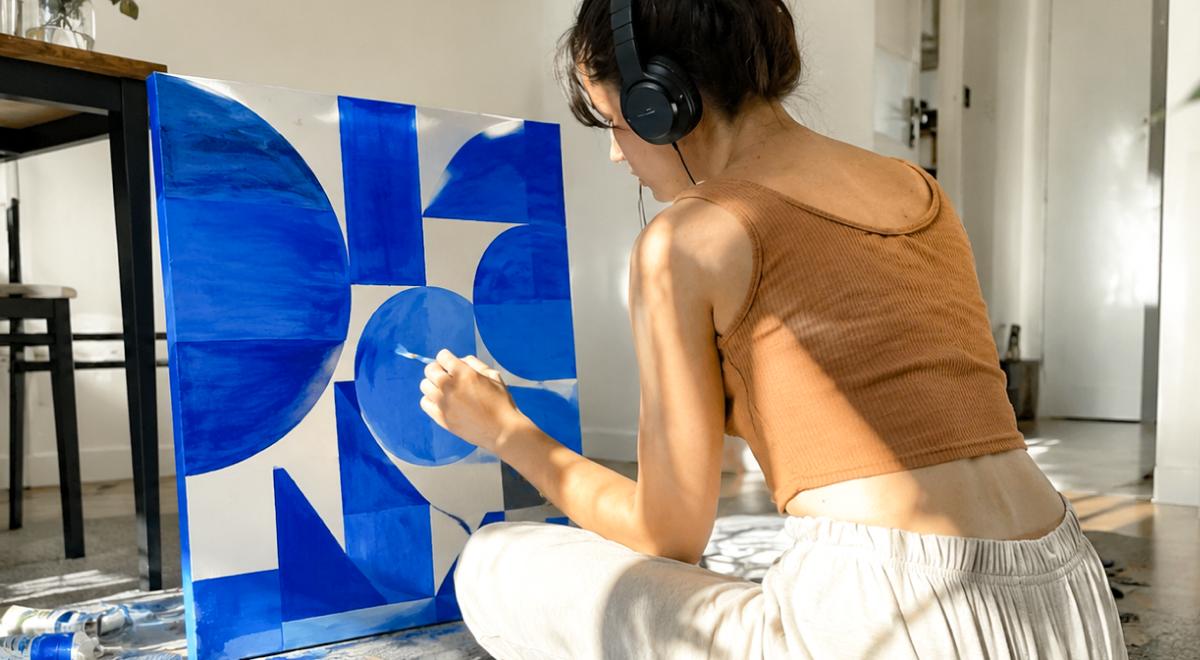 Woman with ADHD wearing headphones and a brown top sitting on the floor at home, painting a blue geometric abstract canvas in a bright sunlit room