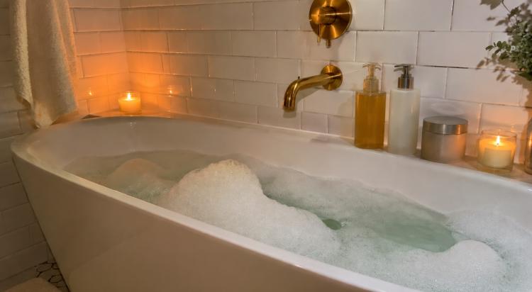 Relaxing night routine bubble bath filling a white tub with a gold faucet, lit candles, soap dispensers, and greenery against white subway tile