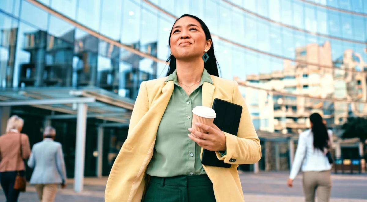 Confident woman in a yellow blazer holding a coffee cup and tablet walking outdoors near a glass office building, representing a career quiz