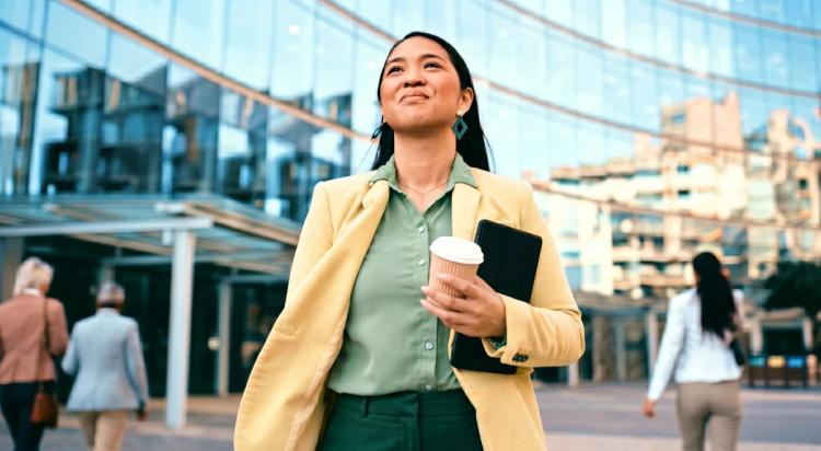 Confident woman in a yellow blazer holding a coffee cup and tablet walking outdoors near a glass office building, representing a career quiz