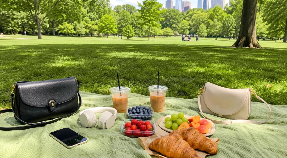 Outdoor picnic spread on a green blanket in a park with croissants, fresh berries, grapes, iced coffee, black and beige handbags, and a phone on a sunny day
