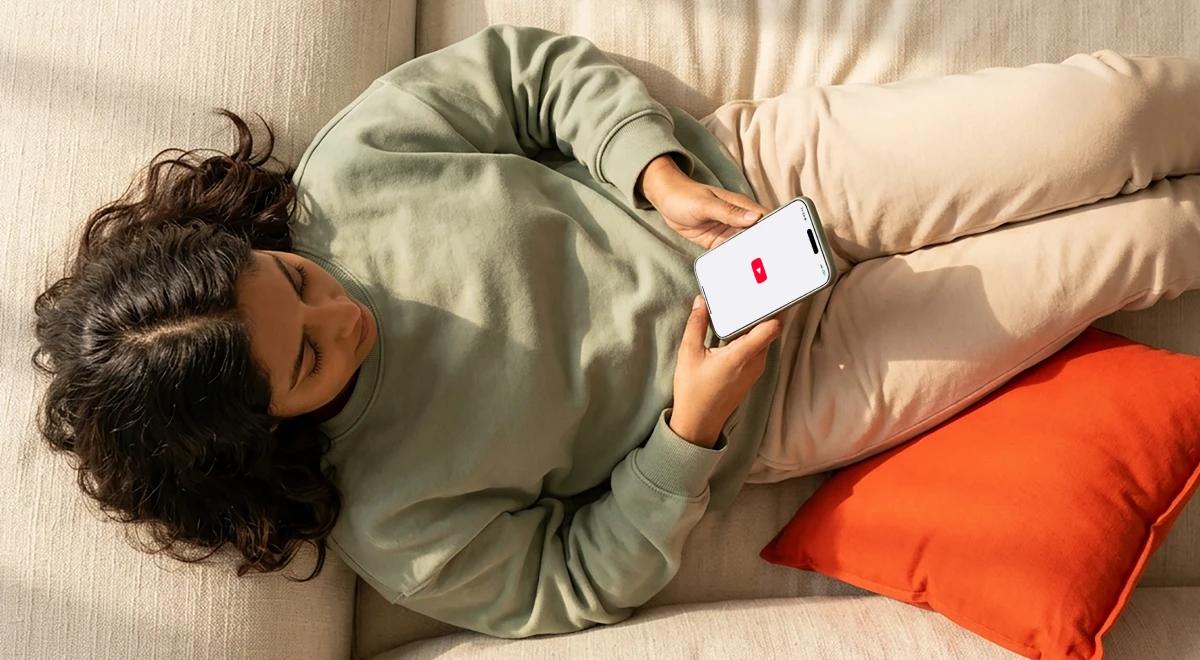 Woman with curly hair lying on a beige couch in a green sweatshirt, scrolling on her phone with a low battery screen, resting on an orange pillow