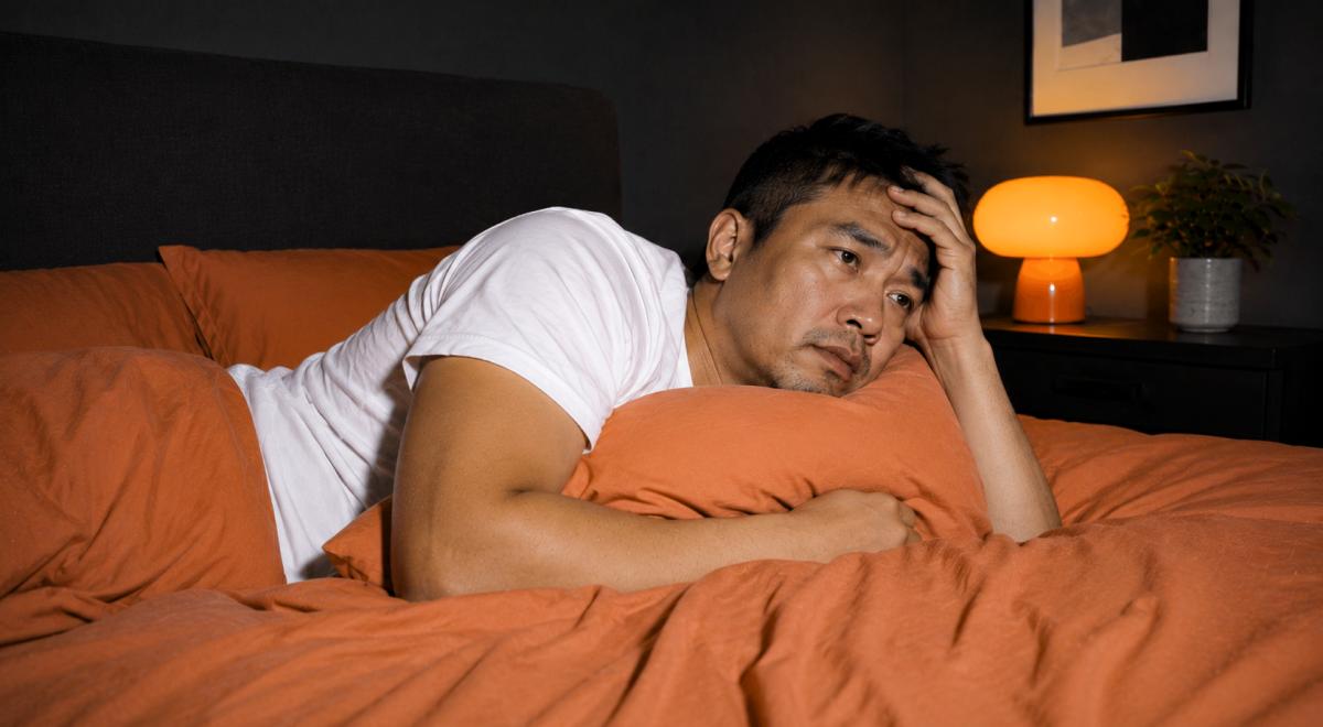 Man lying awake on orange bedding, resting his head on his hand while overthinking at night, with a glowing lamp and plant on the nightstand beside him
