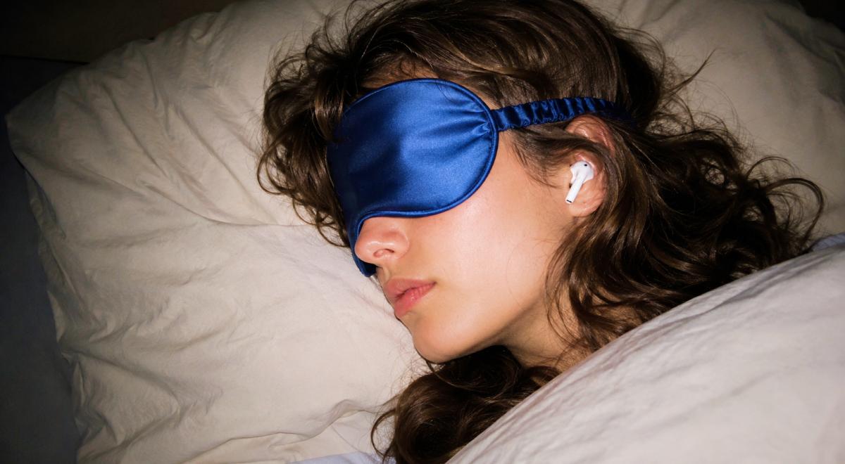 Woman with curly brown hair resting in bed wearing a blue satin sleep mask and a wireless earbud, listening to sleep stories for adults on white pillows