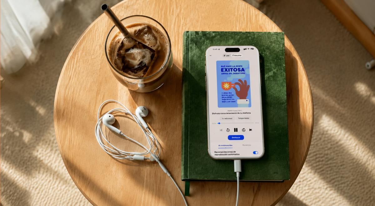 Smartphone sobre un libro verde reproduce un audiolibro para escuchar 20 audiolibros en 20 días, junto a audífonos blancos y un café helado con popote en una mesa de madera