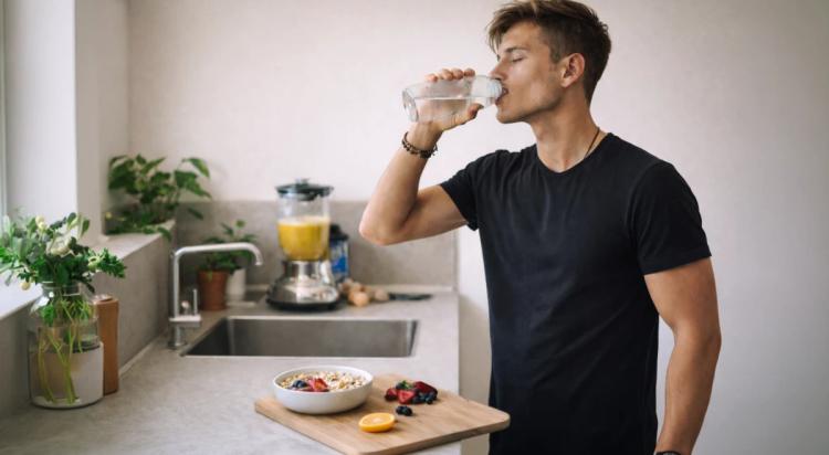 Young man in black t-shirt drinking water in a modern kitchen with a fruit bowl, blender, and orange juice on the counter, inspired by Ashton Hall morning routine and physical health