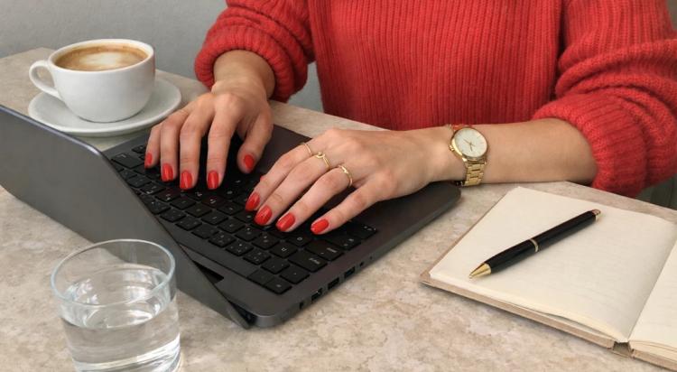 Successful woman in red sweater typing on laptop at marble desk with coffee, water glass, and open notebook as part of her morning routine