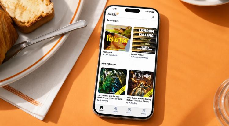 Smartphone displaying the Audible app with bestseller and new release audiobook covers on an orange breakfast table surface with toast and a glass nearby