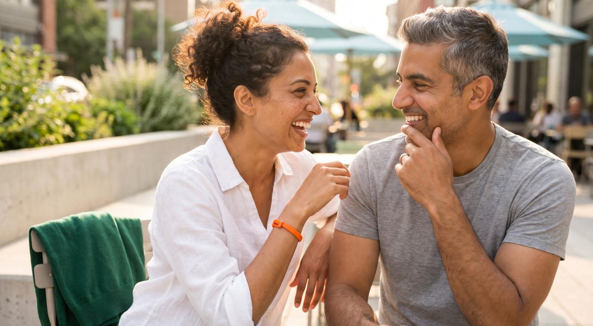 Smiling couple flirting and laughing together at an outdoor city café, the woman in a white shirt and the man in a grey t-shirt gazing at each other in warm sunlight
