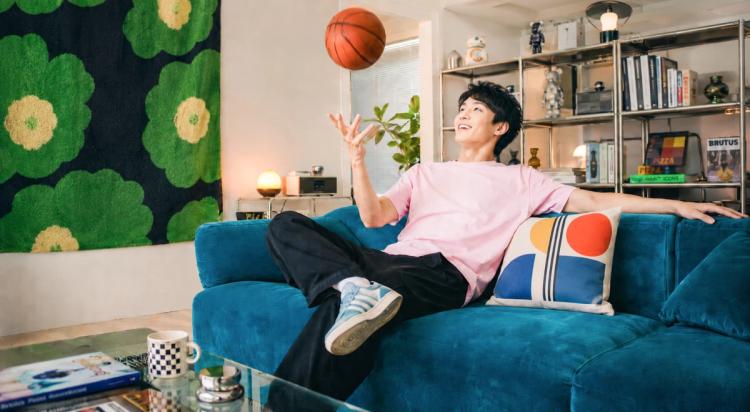 Young Asian man in a pink shirt tossing a basketball while relaxing on a teal sofa in a stylish living room, representing confidence and positive self-esteem