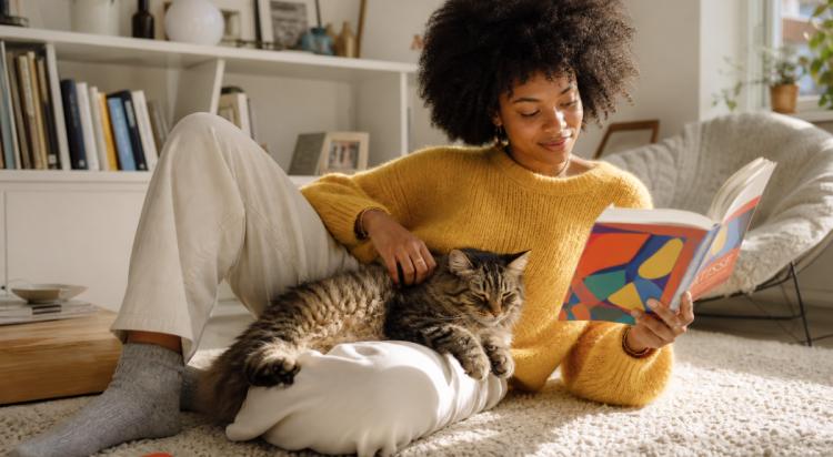 Woman in a yellow sweater relaxing on a cozy living room rug, petting a tabby cat while reading one of her favorite books for creativity beside a bookshelf