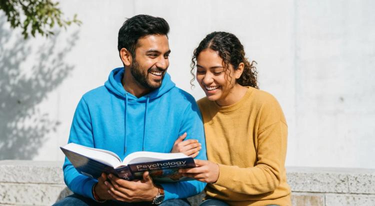 Young man in blue hoodie and woman in yellow sweater sitting outdoors, smiling while reading a Psychology book together against a light concrete wall