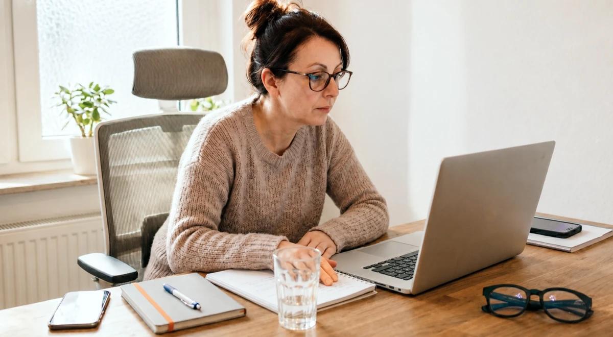 Middle-aged woman with glasses and a beige sweater working on a laptop at a home office desk with a notebook, water glass, and smartphone nearby
