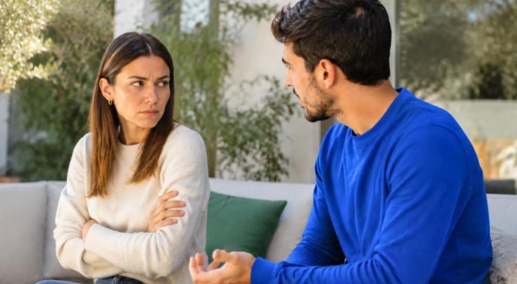 Woman with crossed arms looking away while a man in a blue sweater tries to talk to her on a couch, illustrating anger and conflict in a relationship