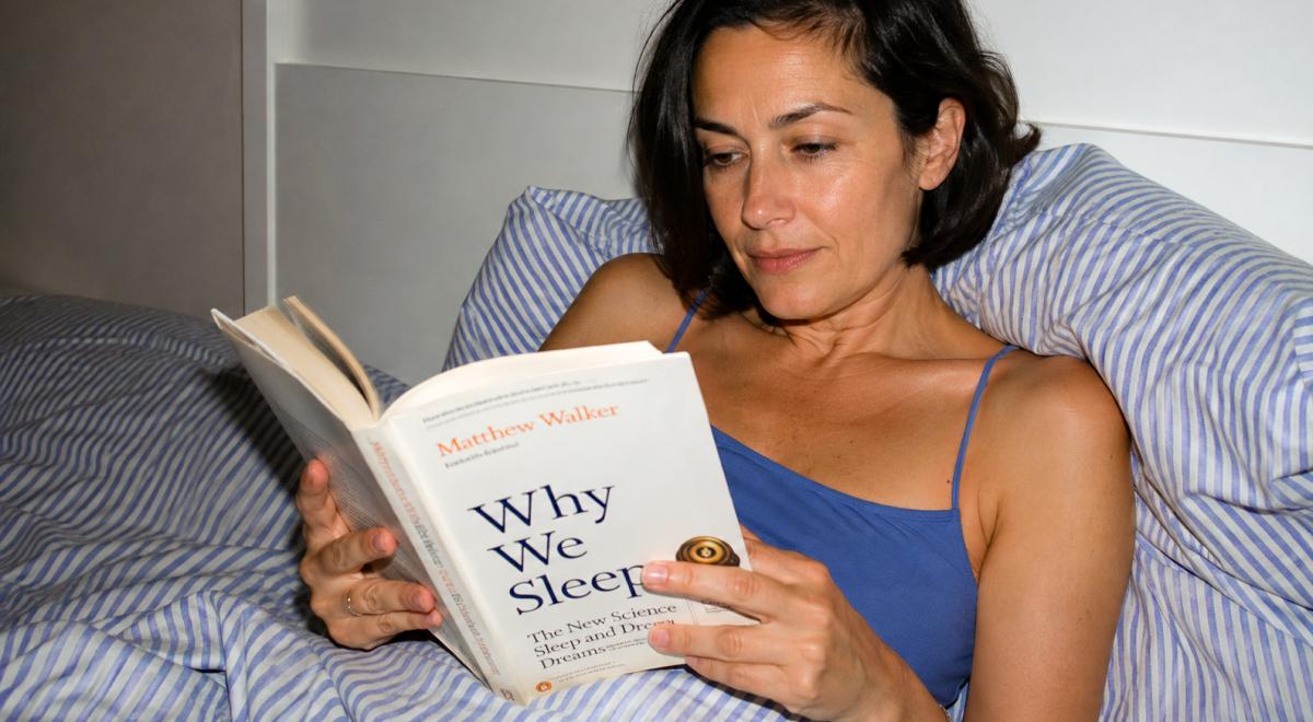 Woman in a blue top relaxing in bed against blue-striped pillows, reading the book Why We Sleep by Matthew Walker before going to sleep