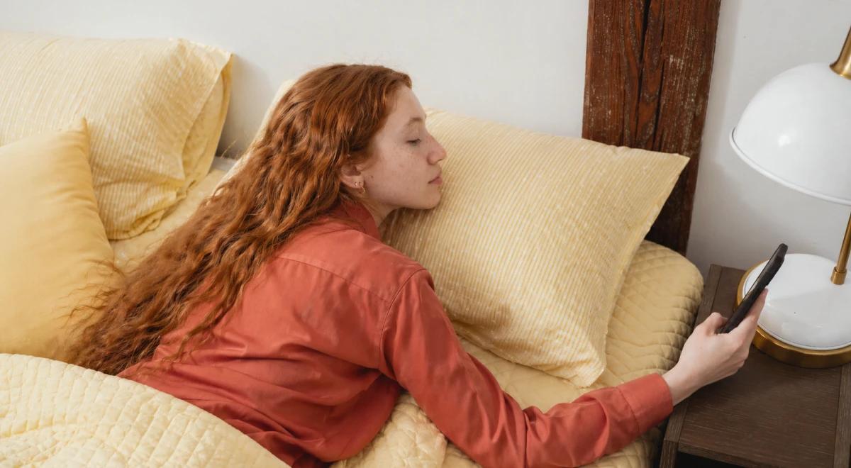 Red-haired woman in a red shirt lying on a yellow bedspread reaching for her phone on a nightstand with a lamp, struggling to wake up early in the morning