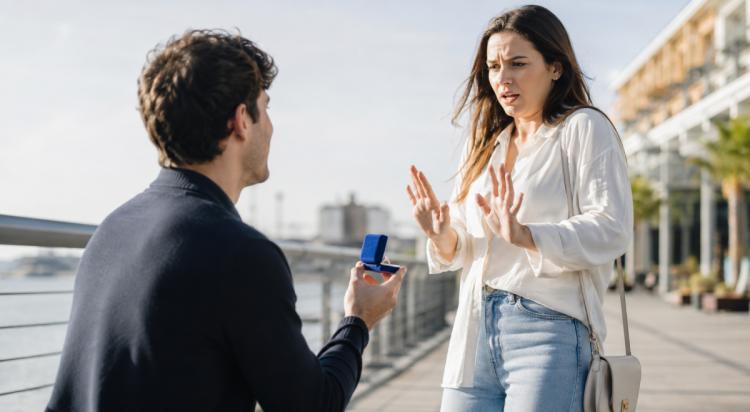 Woman in white shirt and jeans rejecting a marriage proposal from a man holding a blue ring box on a waterfront promenade, illustrating commitment issues in relationships