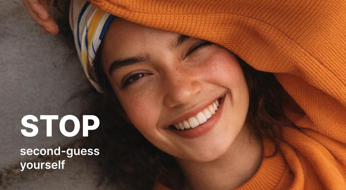 Smiling young woman wrapped in an orange sweater lying on a gray floor with a striped headband, radiating confidence and positive worker mental health free from self-doubt