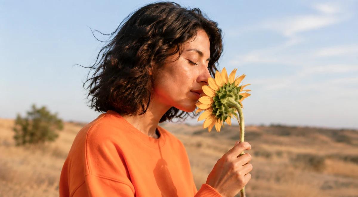 _Young woman in an orange sweatshirt smelling a sunflower outdoors with a dry desert landscape and blue sky in the background