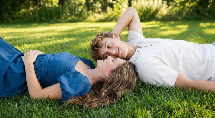 Young couple lying on green grass face to face in a romantic moment, woman in a blue dress and man in a white shirt, with lush green trees in the background
