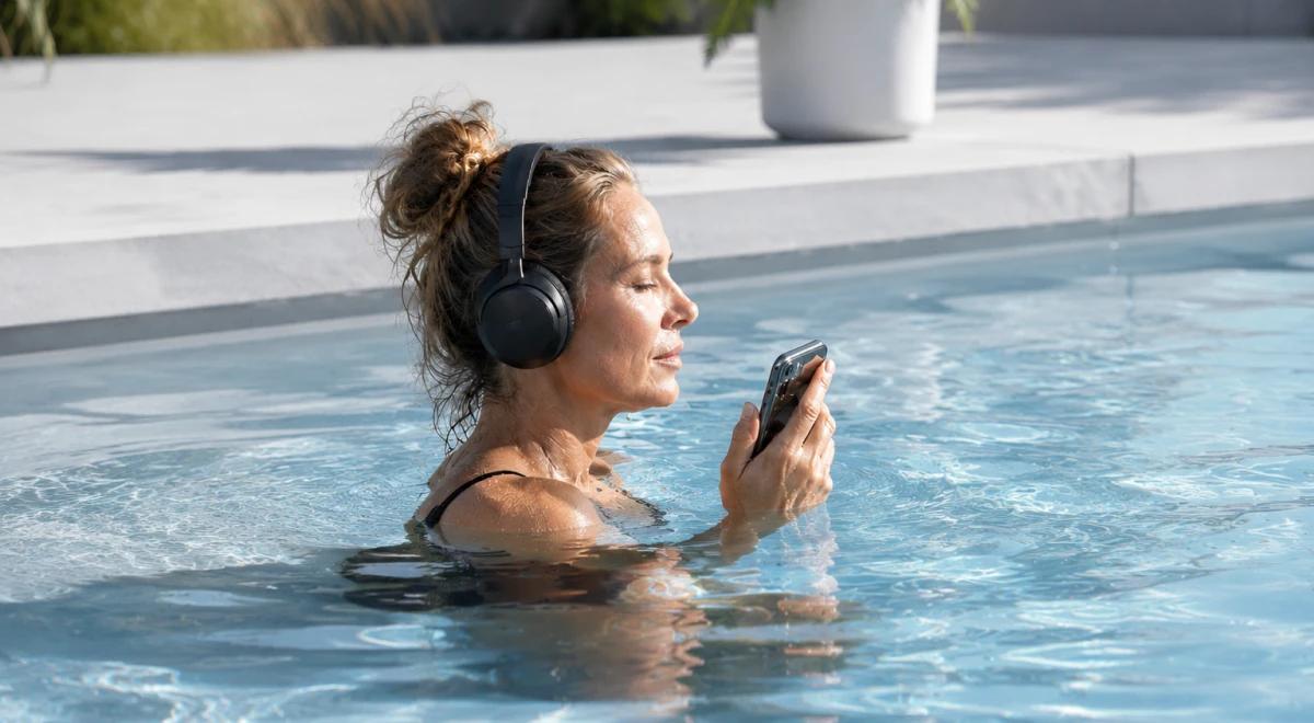 Woman relaxing in an outdoor pool wearing black headphones and holding a smartphone, eyes closed, enjoying a mindful mental reset moment