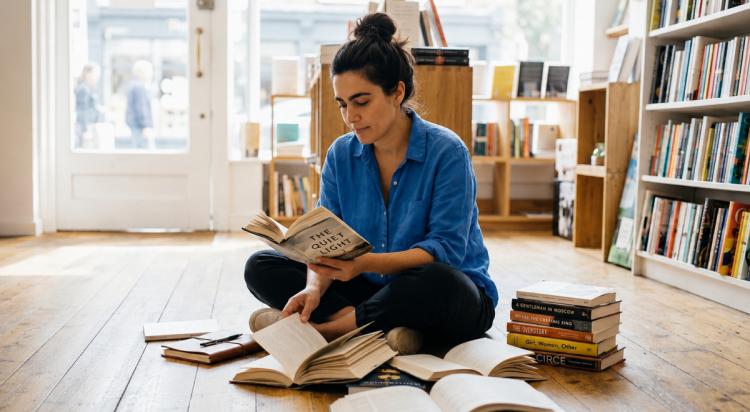 Woman in a blue shirt sitting cross-legged on a wooden floor reading in a bright bookshop, surrounded by open books and stacked reads including the best books 2026 to explore