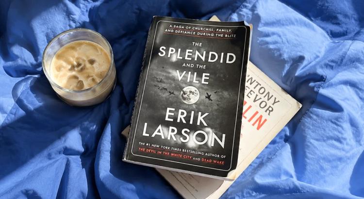 The Splendid and the Vile by Erik Larson placed on top of another book beside an iced coffee drink, all resting on blue fabric bedding (1)