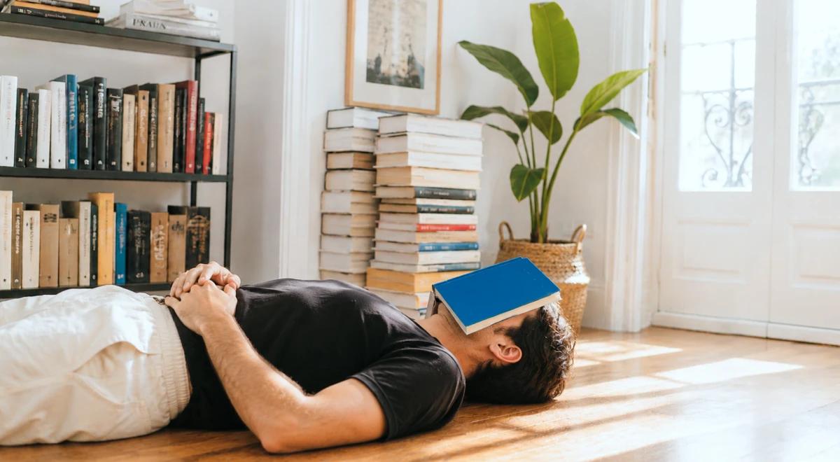 Man in a black shirt lying on the floor with a blue book on his face, surrounded by bookshelves and stacked books in a bright living room
