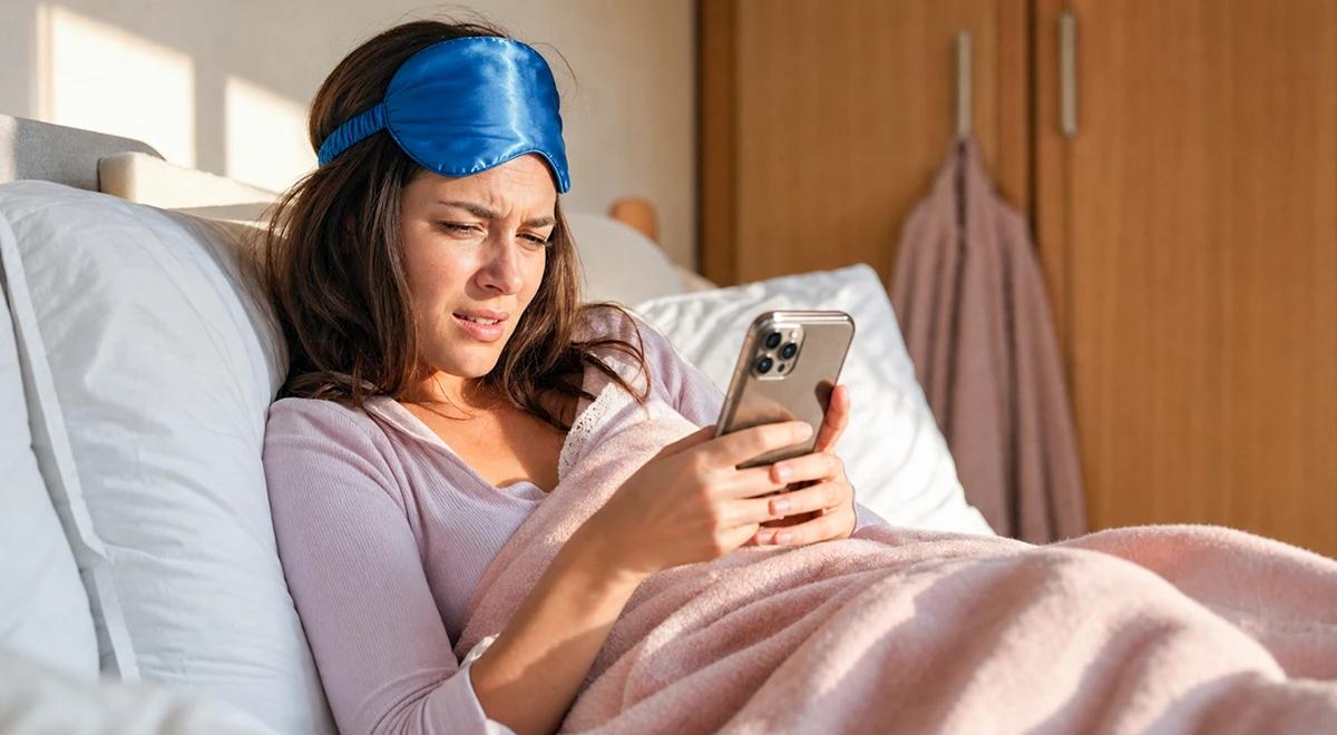 Woman lying in bed with a sleep mask on her forehead, looking upset while checking her phone, illustrating struggles to stop Instagram addiction at night