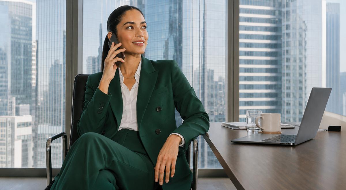 Confident businesswoman in a green suit talks on her phone in a high-rise office, ready to speak like a girlboss, with a laptop, coffee mug and city skyline behind her