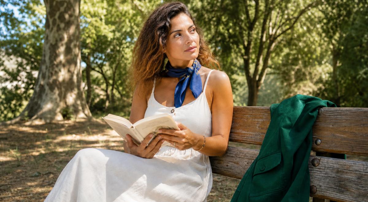 Woman in white sundress and blue neck scarf sitting on a wooden park bench holding an open book, gazing away in a moment of passive reading amid sunlit green trees