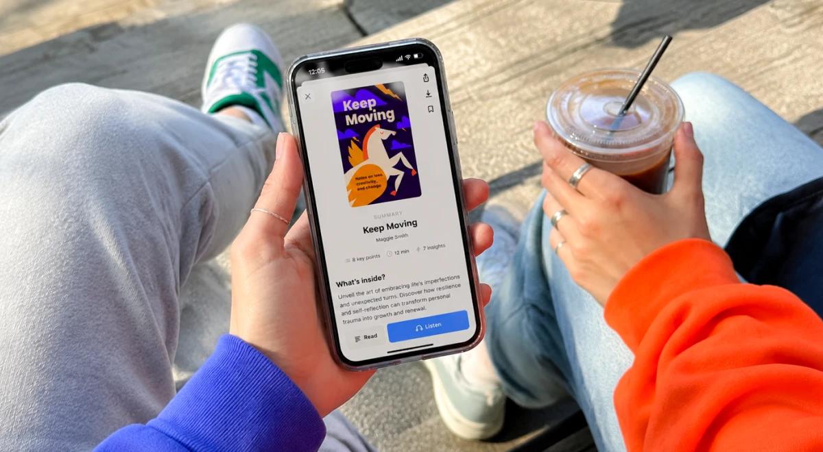 Person in a blue hoodie holding a smartphone with the Keep Moving app open, sitting outdoors with an iced coffee drink, wearing green sneakers
