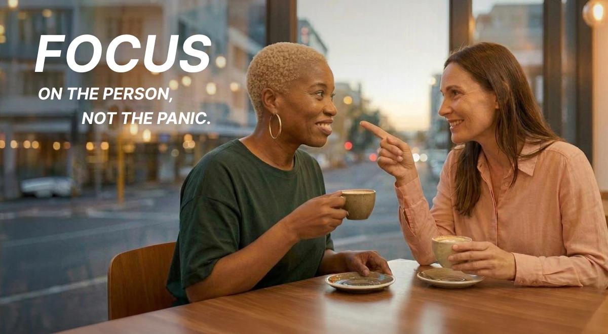 Two women sharing a warm conversation over coffee at a café with a blurred city street at dusk in the background, with a bold text overlay about focusing on the person not the panic