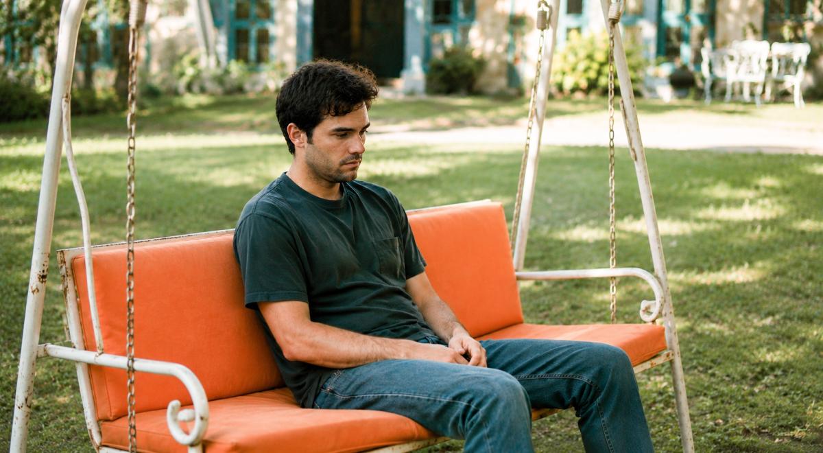 Sad single man in a dark t-shirt and jeans sitting alone on an orange-cushioned garden swing on a green lawn outside a house
