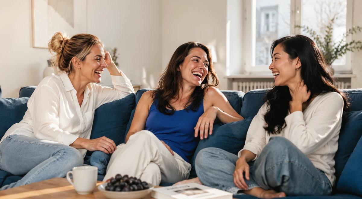 Three happy women laughing together on a navy sofa in a bright living room, enjoying each other's company