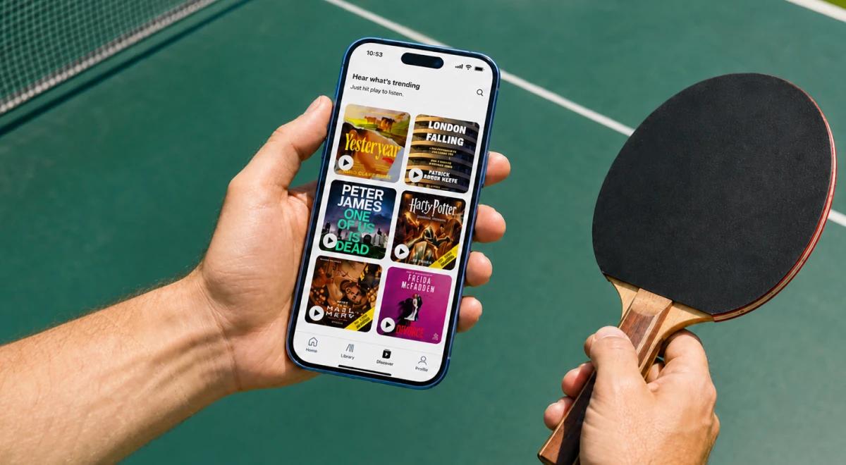 Man holding a smartphone displaying an audiobook app with trending titles and a ping-pong paddle, standing at a green table tennis table