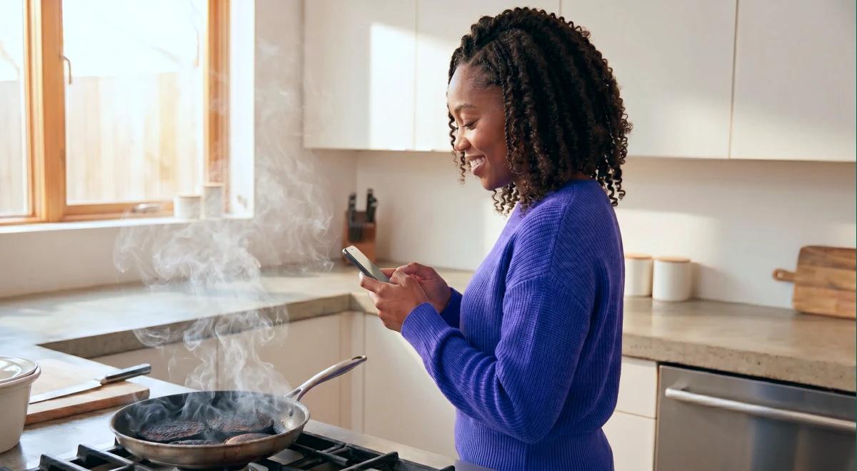 Black woman in a purple sweater smiling at her phone while food burns in a smoking pan on the stove, illustrating a short attention span and distraction in a bright modern kitchen