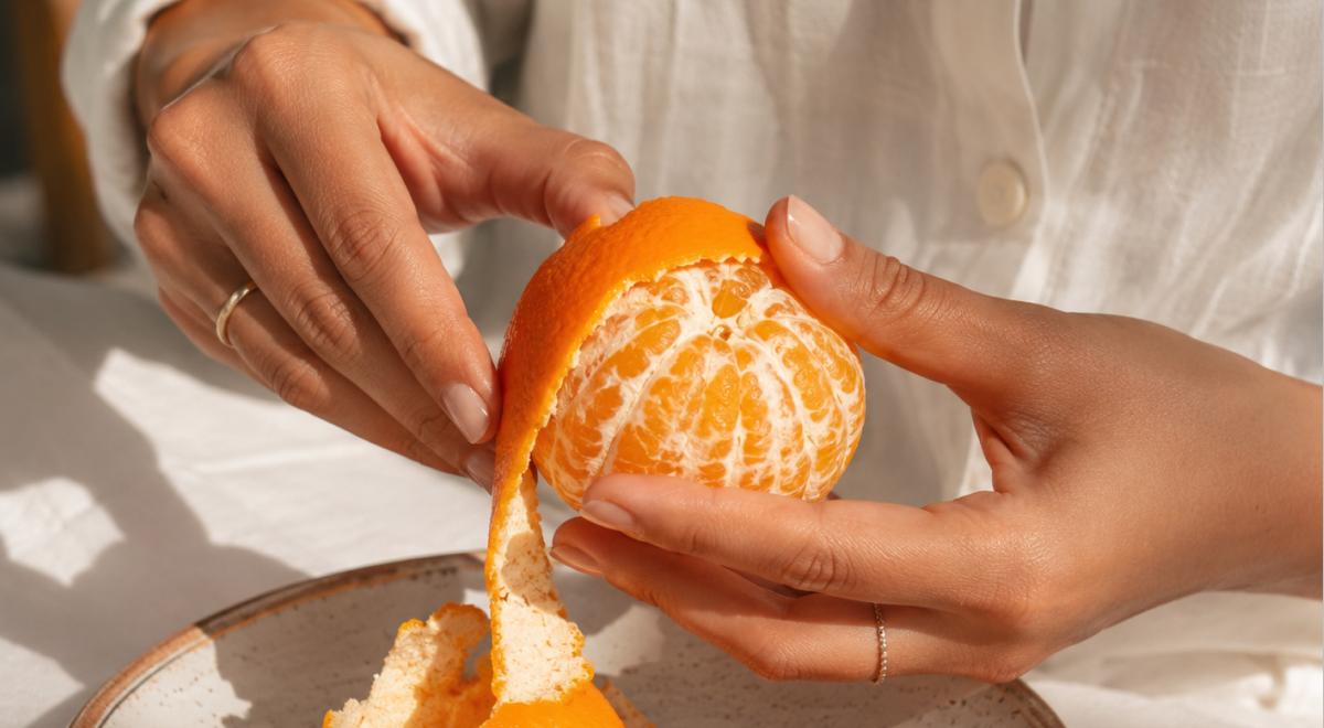 Woman in a white shirt peeling a fresh orange over a speckled ceramic plate in soft natural light, illustrating the orange peel theory of small caring gestures in relationships