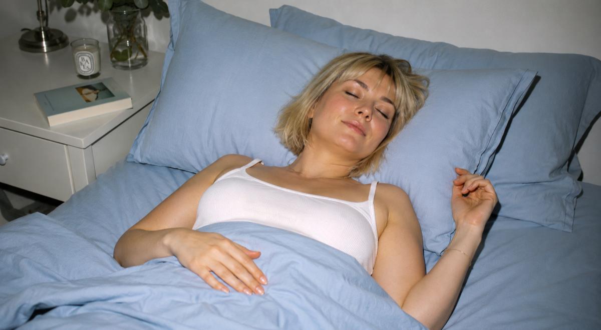 Blonde woman peacefully sleeping in a bed with blue linens, showing how to quiet your body to sleep, beside a nightstand with a book and candle