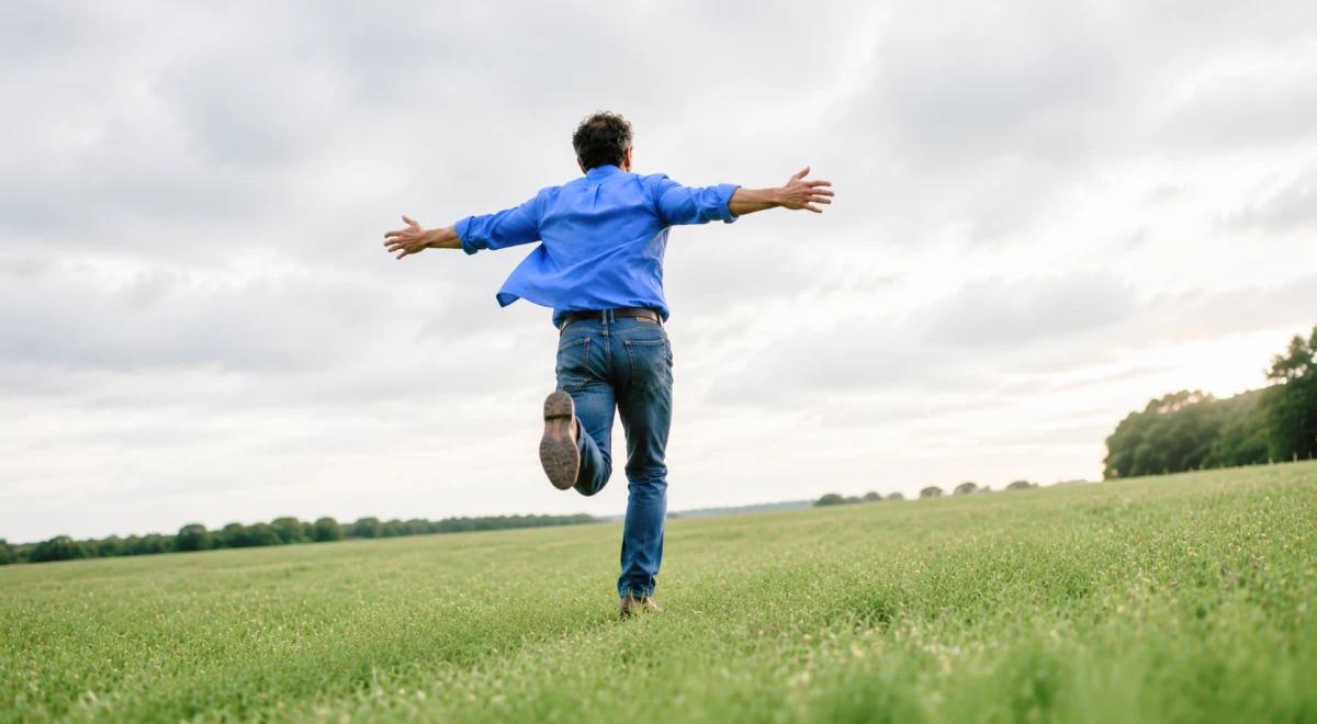 Person in a blue shirt running freely across an open green field with arms spread wide, symbolizing freedom and secure attachment