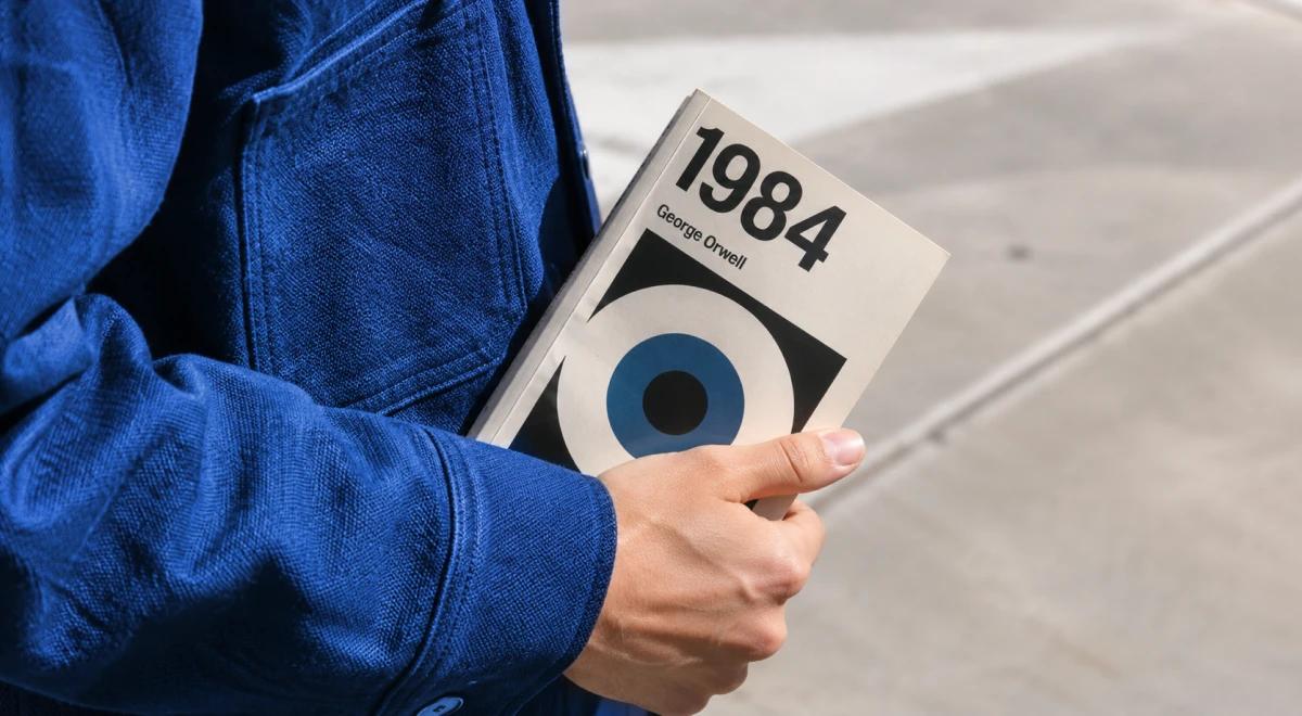 Person in a blue jacket holding a copy of 1984 by George Orwell outdoors on a concrete surface, featured among must-read books before you die