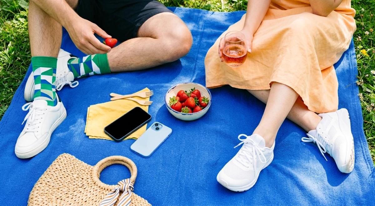 Two people sitting on a blue picnic blanket outdoors, sharing strawberries and rosé wine, with a smartphone, wicker bag, and yellow napkins on green grass background