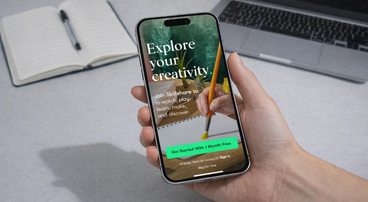 Hand holding a smartphone with the Skillshare app on screen, notebook and laptop in background, highlighting the platform's microlearning subscription cost offer