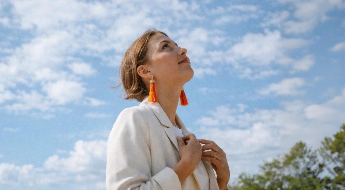 Woman in a white blazer with orange tassel earrings looking up at a blue cloudy sky, embracing a growth mindset and building a habit of self-reflection outdoors