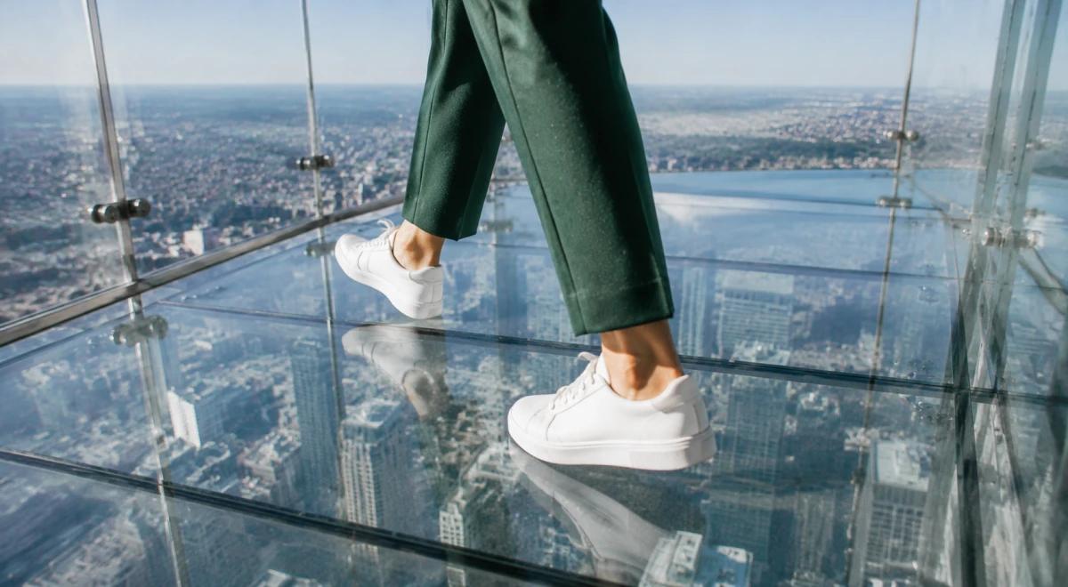 Woman in a green blazer and white sneakers sitting on a glass-floor observation deck with a panoramic city skyline view below, representing fearlessness and motivation
