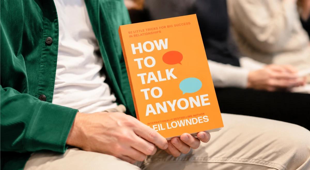 Person in a green jacket holding the orange How to Talk to Anyone book by Leil Lowndes, with other people blurred in the background